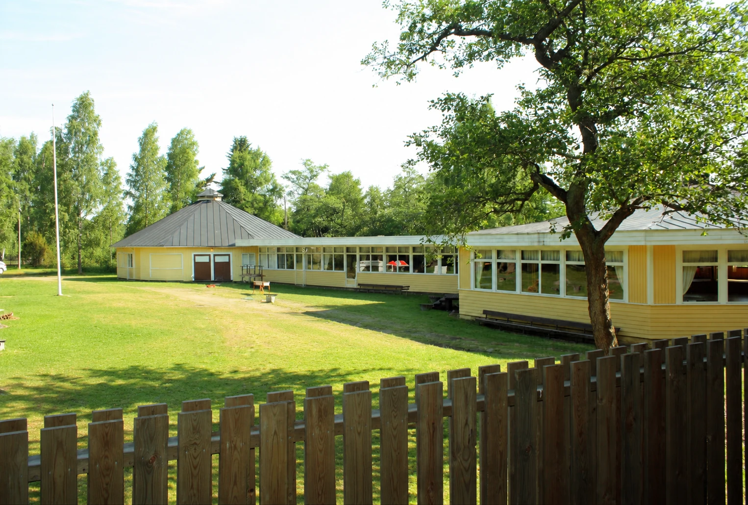 Rörgrunds paviljong med en solbelyst gård, stora träd och trästaket.